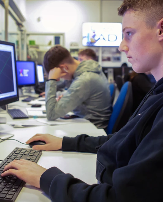 A student in a dark hoodie concentrating on a dual-monitor CAD workstation, modeling a 3D mechanical part. A student in a dark hoodie concentrating on a dual-monitor CAD workstation, modeling a 3D mechanical part.